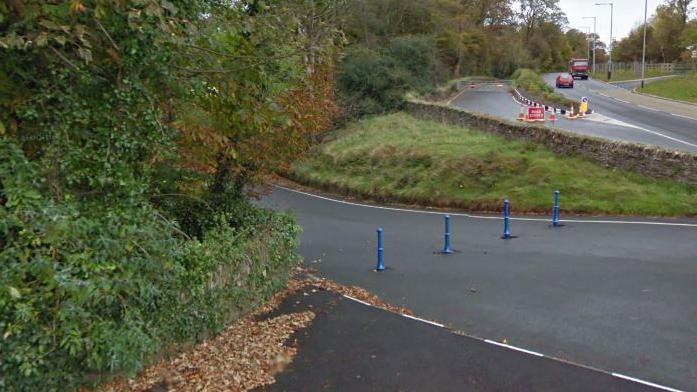 The Governor's Dip road coming out at the roundabout at Governor's Road. there are trees and bushes to the left and blue traffic bollards across the road.