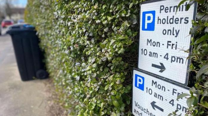 Parking permit sign - describing restrictions - nestled into green hedge, with a wheeled bin on the street in the background.