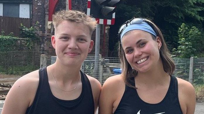 Izzy Flynn and Maisie James smiling in black gym clothes on a sunny day in front of a railway bridge