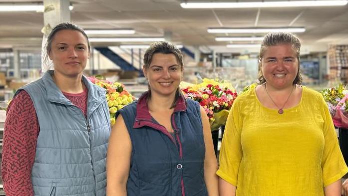 Four people stand in a row in a flower packing warehouse. Three women from left to right wearing a blue vest, a navy vest, bright yellow top, and a man in a navy polo at the end. The flowers behind them are wrapped in yellow paper, and coloured pink, white and red. 