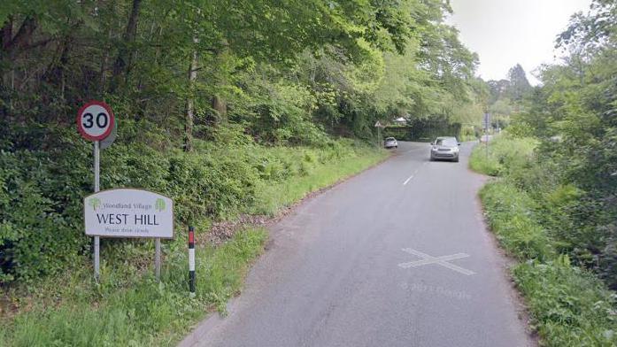 A white sign welcoming visitors to the village of West Hill. It is on the side of a narrow road with grass and trees either side of the road. A grey car is driving along the road. 