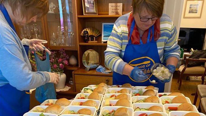 Two women wearing blue aprons with the charity's gold logo prepare meals of bread rolls and salads on a table. In the background is a cabinet containing glasses, photographs and ornaments. 