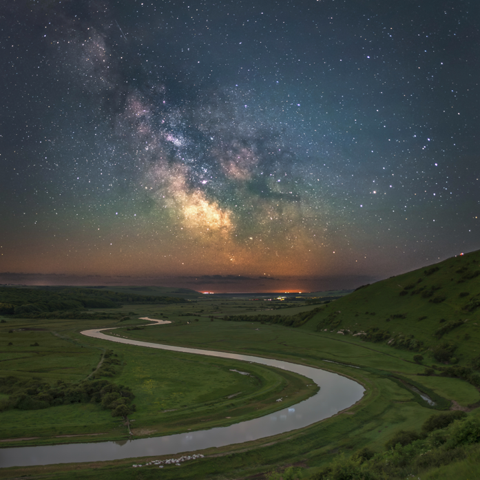 The Milky Way can be seen in a colourful night sky. The image shows a winding road snaking through a landscape of green. 