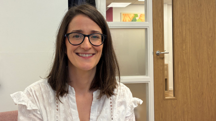 Leanda Lee, smiling at the camera has dark shoulder length hair, blue eyes and is wearing dark framed glasses and a white blouse.