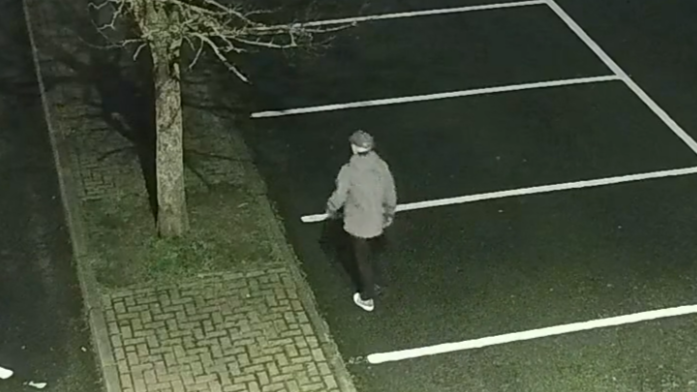 A CCTV image of a man walking through a car park wearing dark trousers, a grey jacket, trainers and a hat.