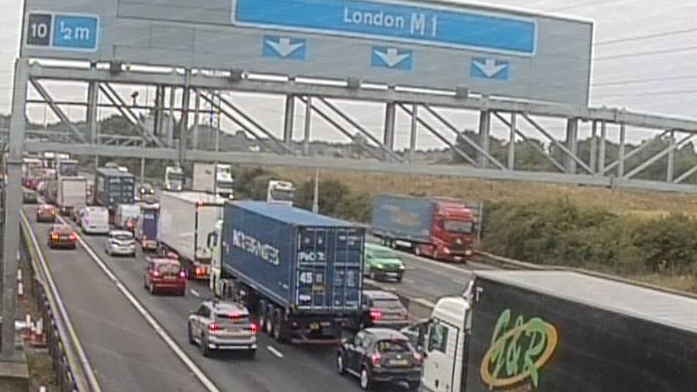 View of a motorway from the left side. Traffic is queuing on the left side of the motorway and on the other side it is flowing. A sign goes above the road that says London M1, with arrows pointing to three lanes.