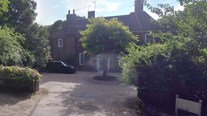 A large red-bricked house with three chimney stacks is obscured by a tree in a front courtyard. A wide driveway leads toward the courtyard, with foliage on either side. 