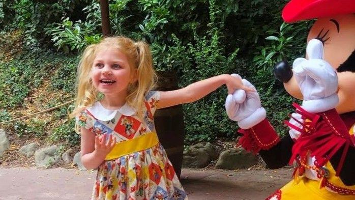 Beatrice, with blonde pigtails, smiling and holding out her hand to Mickey Mouse at Disneyland. She is wearing a patchwork dress featuring Toy Story characters.