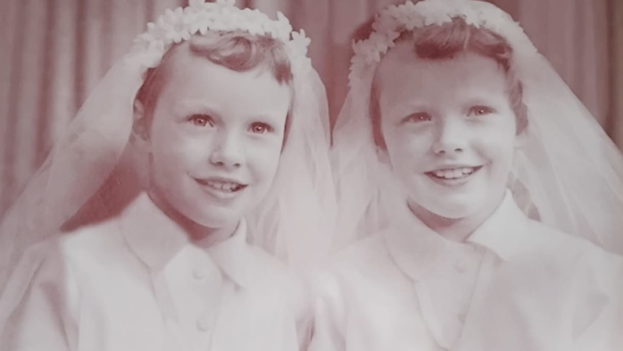 Black and white image of Geraldine and her twin sister Elizabeth as children. Both girls are smiling at the camera while standing next to each other. They are almost identical. They are wearing white collar blouses and veils with flowers atop their hair. 