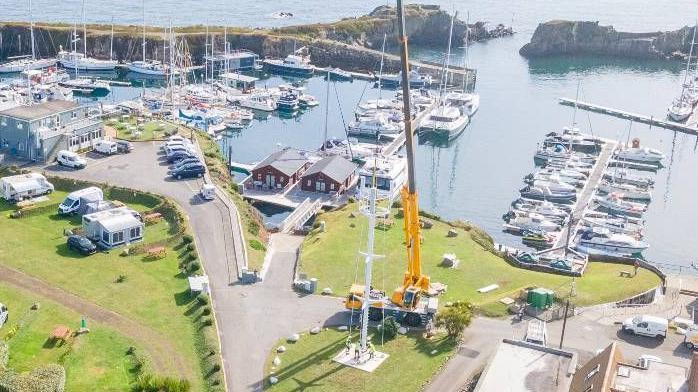 The new mast which is white stood in the middle of a piece of green land, a yellow crane is next to it seeming to install the mast 