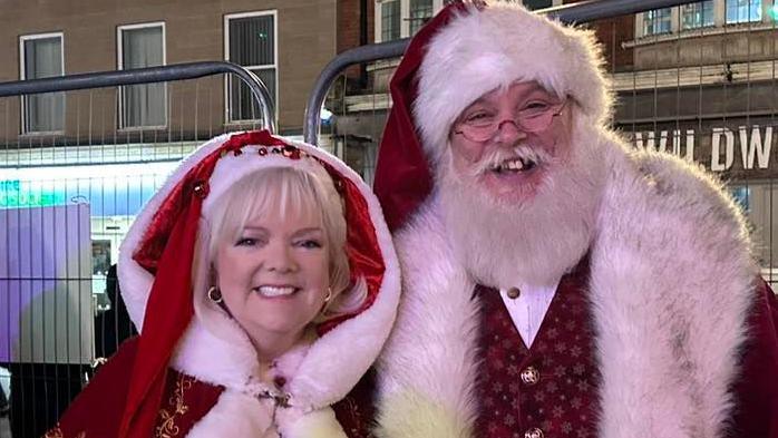 A Father Christmas dressed in red suit with white beard standing next to Mrs Claus, also in red suit with hat and cap and white hair. They are both smiling and looking straight at the camera. They are in front of a fence with businesses behind