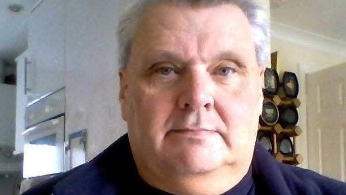 A head shot of a man in his home, he has grey hair and is wearing a blue top.