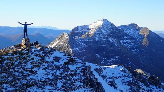 A walker stands on top of a cairn in the Torridon mountains. The impressive peaks rise behind them and the sky is blue.