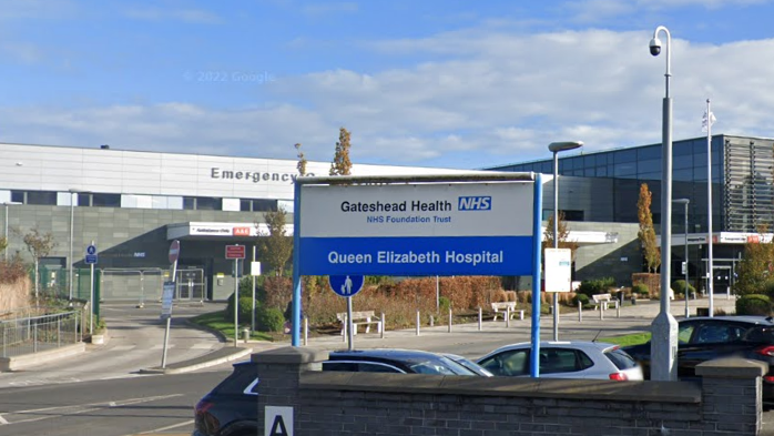 The outside of the Queen Elizabeth Hospital, which is marked with a blue and white sign. Behind is a large, grey building with a car park in the foreground. 