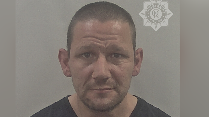 A police custody photo of John Atherton looking at the camera. He has short shaven black hair and short dark stubble on his face. He is wearing a black t-shirt. The wall behind him is grey.