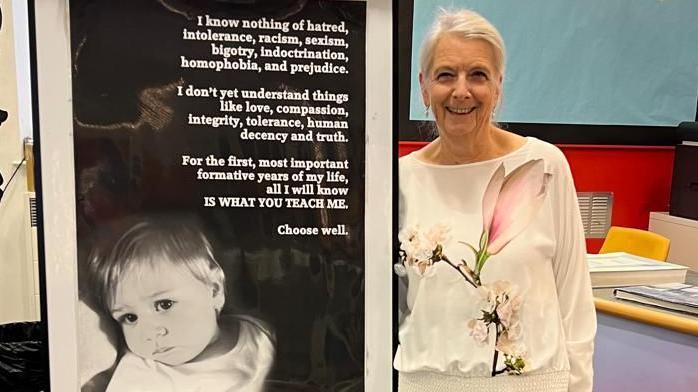 Jennie Thompson, who has grey hair tied back and is wearing a white top with an elaborate floral design in the front. She is standing next to a poster featuring a black and white photo of a young child alongside text.