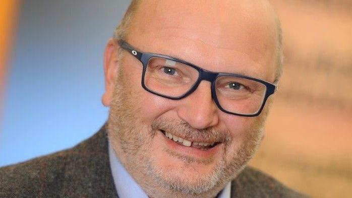 A man with a grey beard and glasses smiles at the camera, pictured in a close-up to his face. He is wearing a tweed suit jacket and shirt.