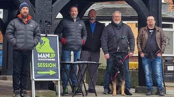 Five men stand on the pavement. One of them stands next to a sign which says MAN UP Gloucestershire session with an arrow beneath the words pointing to the right. One of the men has a dog on a lead.