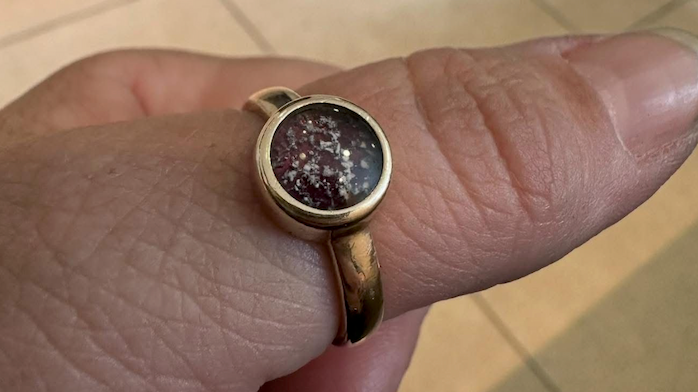 Mrs Russell's hand with a gold ring on her thumb.  It is gold and has a circular cut face which is a deep red wine colour. On top are fragments of her daughter's ashes fused to the ring.