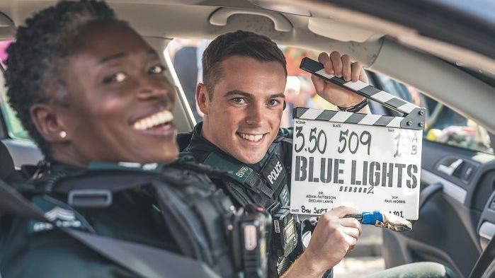 Two cast members of the series Blue Lights, one woman and one man in PSNI uniforms in a police car. The man is holding a Blue Lights clapper board, both are smiling     