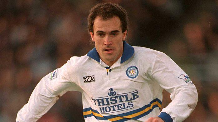 A male footballer with short brown hair runs with his tongue sticking out his mouth. He is wearing a white, blue and yellow Leeds United shirt.