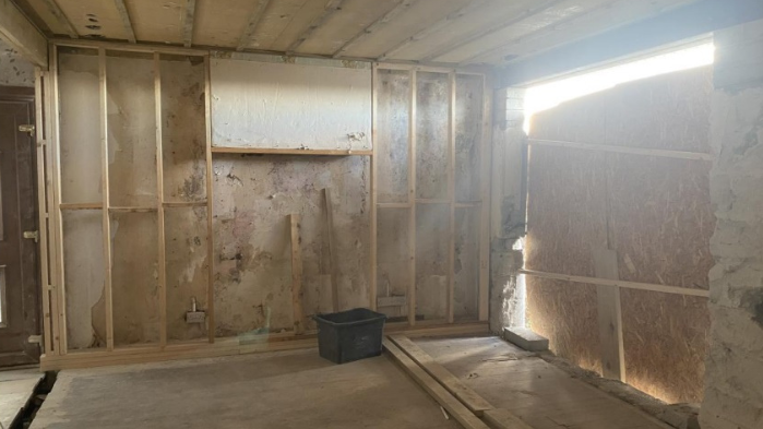 A shell of a room with exposed timber beams and chipboard loosely secured against a large rectangular hole in a breeze block wall.