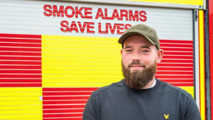Matt Welsh in a navy Lyle and Scott jumper and a khaki coloured baseball cap poses for a photo by the side of a fire engine.