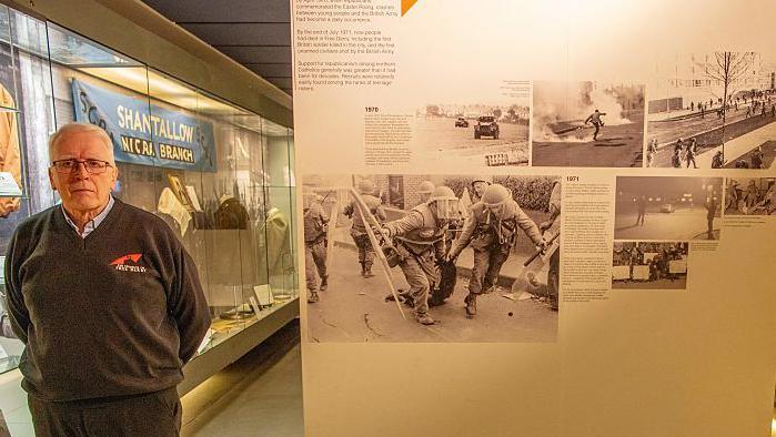 John Kelly has short grey hair, black metal glasses and is wearing a grey jumper with a blue shirt and grey trousers as he stands beside a wall in the museum with black and white images on it.