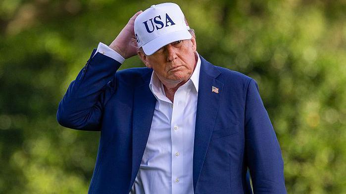 US President Donald Trump wearing a blue suit and a white cap with USA on it