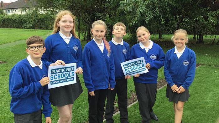 Six children wearing their Uphill Village Academy uniform, which consists of blue cardigans and black trousers or skirts. They are standing on the grass at their school with trees behind them. They are holding two signs which say 'Plastic Free Schools' and smiling at the camera.