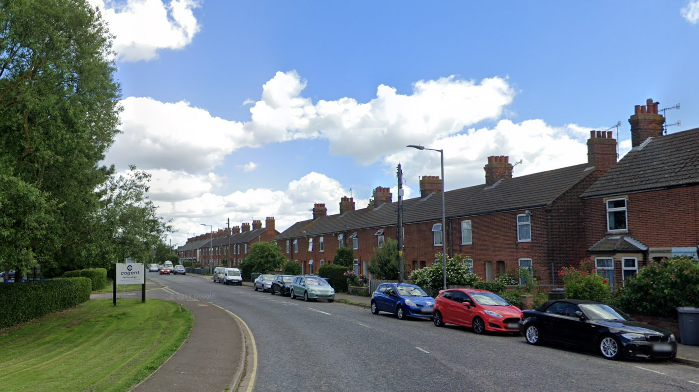 A residential street including a row of red-brick houses, with a line of cars parked outside. On the other side of the road is a green space. 