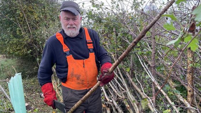 A man with a grey beard and moustache looking to the camera holding a large branch in his left hand and a machete-style knife in the other. He has a bright orange overall on and is wearing red gloves and a blue cap. He is stood next to a row of hedges.