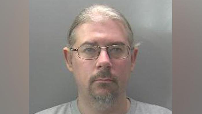 Robert Norman is looking at the camera in a police custody shot. He has fair hair that appears to be tied back, and a beard and moustache. He is wearing spectacles and a grey top.