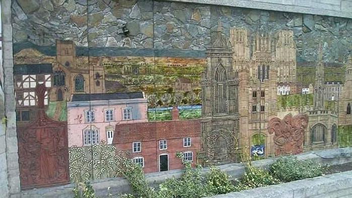 A mural in muted shades including red, pink and stone, showing various buildings including houses and Wells Cathedral.
