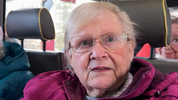 Barbara Denison, is sitting on a bus and has grey hair and glasses. She's wearing a purple top and a white jumper.