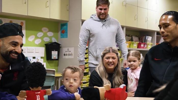 Isaac clutch his LFC plush toys while sitting at a class table with his mum holding his big sister and dad behind him. Mohamed Salah sits to the left with Virgil van Dijk sits on the right