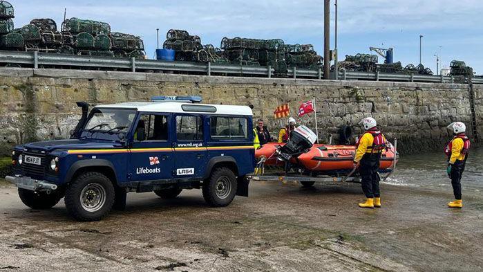 Small lifeboat attached to 4X4 car surrounded by people in rescue gear