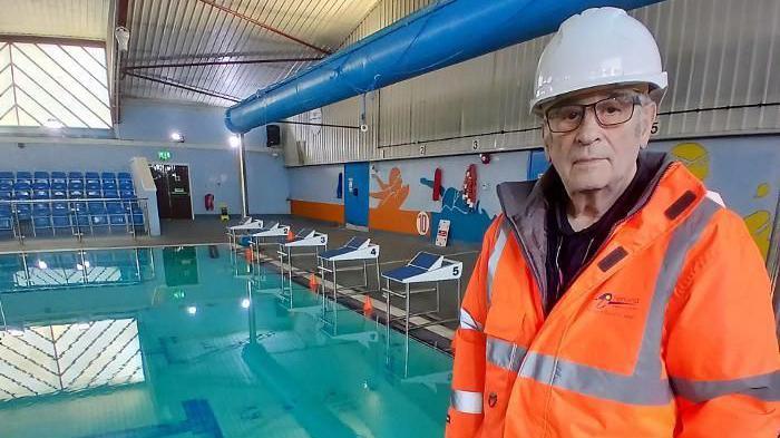 Image shows swimming pool with seats to one side. Councillor Alex Miscandlon appears in an orange hi-vis jacket and white hard hat in the foreground of the picture. 