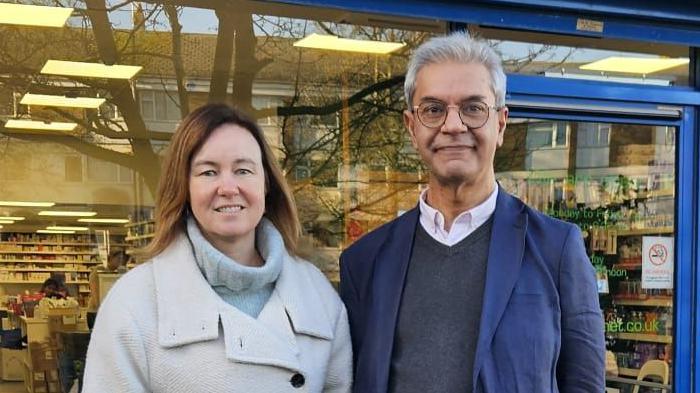 Marie Goldman and Dipak Pau wearing coats outside a pharmacy in Essex. They are both smiling at the camera