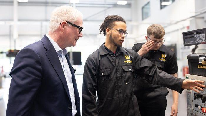 A man with white hair and glasses, wearing a suit, watches as a student in black overalls points towards some equipment. Another student stands in the background