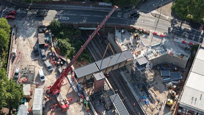An aerial shot of a large crane placing a footbridge above a train track