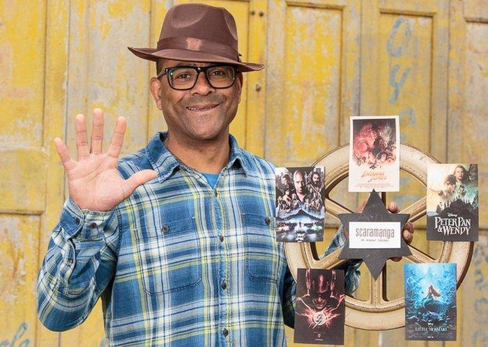 Carl wears an Indiana Jones style hat holding a cut out film canister with five film posters which feature items from the shop