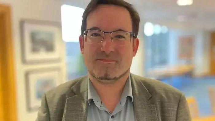 Lawrie Hooper, who has short brown hair and a thin goatee beard and is wearing glasses, a light coloured suit jacket and a grey open-necked shirt.