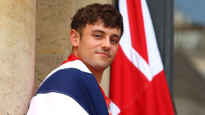 Tom Daley smiling with a British flag around his shoulders and in the background.