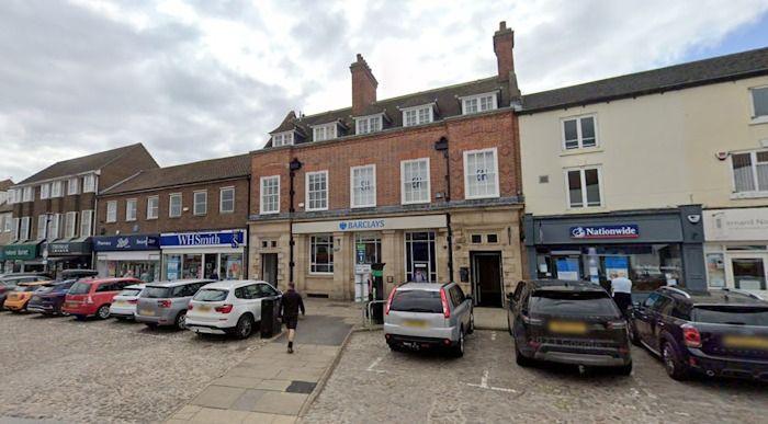 A cobbled square with cars parked on it and a lage brick building in the centre of the image in a mixture of white and red brick with Barclays bank signage