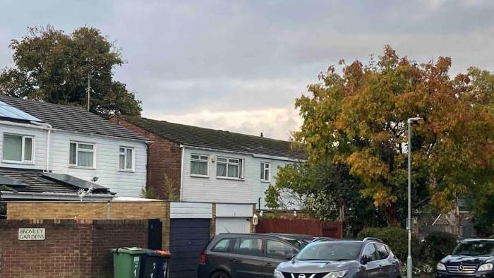 Bromley Gardens: A square black-and-white sign on a red bricked wall. Behind the wall is a line of red-bricked houses with white cladding. A green rubbish big and a grey rubbish bin stand outside the houses next to several parked cars.