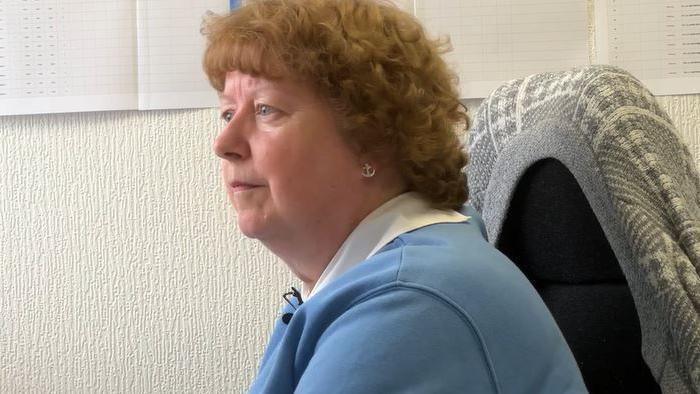 A woman with light red hair in a perm is sat at her desk working. She is wearing a blue jumper over a white shirt and has a grey cardigan over the back of her chair. Behind her on the wall are office planners and charts.