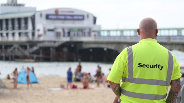 A man wearing a yellow high vis top with security on the back. He is stood on a beach in front of a pier.