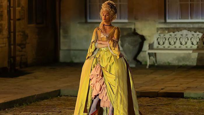 Actress Marisa Abela, as Yasmin in Industry. She is pictured wearing period costume - a yellow ballroom gown from the 18th century French court, and a blonde wig. She stands with her arms crossed, with the sandstone walls of a stately home behind her.
