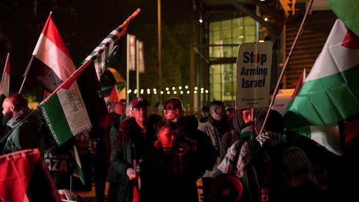Pro-Palestinian campaigners stage a demo outside Villa Park. Many are carrying flags and waving them. There is a banner that says "stop arming Israel". The stadium building is behind them with the lights on inside.